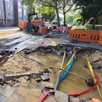 Im Asphalt auf einem Bürgersteig ist ein großes, mit Wasser gefülltes Loch zu sehen. Rohre verlaufen durch das Loch. Drumherum stehen Bauzäune und im Hintergrund sind Bagger und Arbeiter zu erkennen.