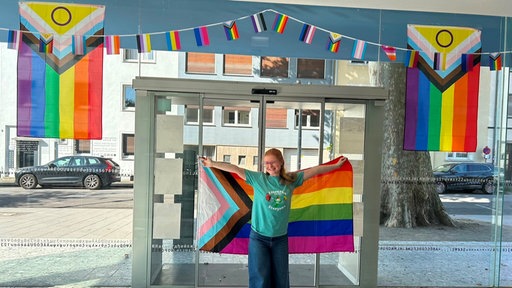Am Eingang der Mönchengladbacher Zentralbibliothek hängen Regenbogen-Gilanden. Lyssa kahlert, eine Mitarbeiterin der Bibliothek, steht davor und hält eine Regenbogen-Flagge.