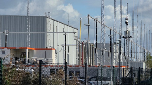 Nahaufnahme der Konverteranlage in Osterath. Auf der rechten Bildhälfte stehen Strommasten, auf der linken Hälfte ist eine graue Halle zu sehen.