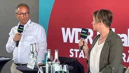 Ein Mann und eine Frau jeweils mit Mikro an zwei Stehtischen schauen sich an