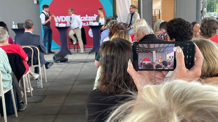 Eine Zuschauer macht ein Handyfoto, im Hintergrund ein Podium mit drei Gästen und einem Moderator
