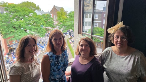 Vier Frauen stehen am Fenster und lachen in die Kamera. 