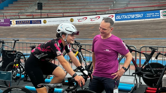Eine junge Frau sitzt auf einem Fahrrad, sie trägt einen weißen Helm und ein schwarz-lila Trikot, neben ihr steht ein Mann mit einem lila T-Shirt, im Hintergrund sieht man eine hölzerne Bahn.