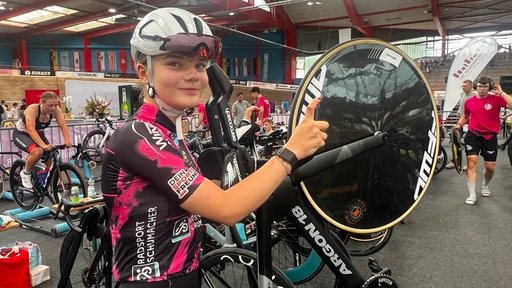 Eine junge Frau sitzt auf einem Fahrrad, sie trägt einen weißen Helm und ein schwarz-lila Trikot, im Hintergrund sieht viele Fahrräder.