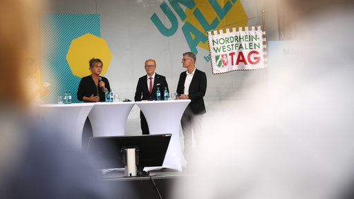 Mona Neubaur, Tilmann Fuchs und Andre Kuper stehen auf einer Bühne.