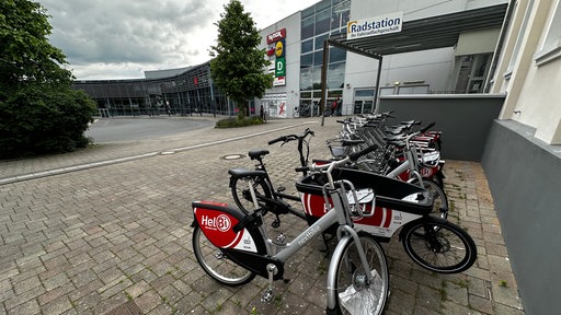 Fahrradverleihstation mit mehreren silber-schwarzen Mieträdern der Marke "nextbike" mit dem roten Logo "HeiBi" vor einem modernen Gebäude mit Glassfassade.