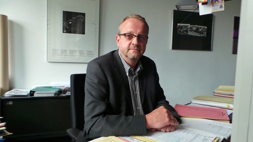 Ein Herr mit Brille und Anzug sitzt in einem Büro und blickt in die Kamera.