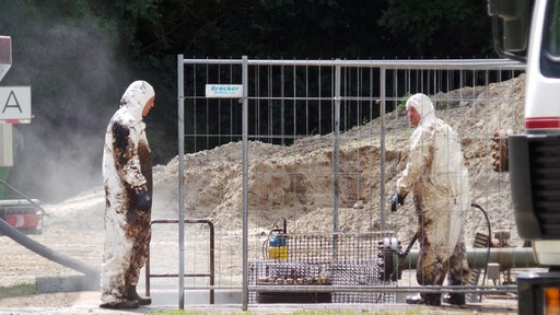 Zwei Männer in weißen Schutzanzügen stehen an einer Kaverne. Ihre Anzüge sind mit braunem Öl besudelt.