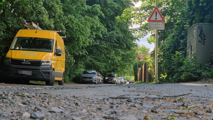 Eine Straße mit Schäden, ein Straßenschild weißt daraufhin. Am Rand sind parkende Autos zu sehen.