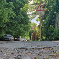 Eine Straße mit Schäden, ein Straßenschild weißt daraufhin. Am Rand sind parkende Autos zu sehen.
