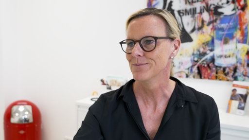 Ärztin Dr. Karin Heinen, eine Frau mit schwarzer Bluse und schwarzer Brille, sitzt an ihrem schreibtisch, hinter ihr bunte Bilder.