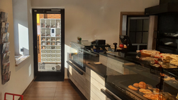 Ein Raum mit einer Theke, in der sich Backwaren befinden. Zudem sieht man einen Brotautomaten, der draußen vor der Tür steht.