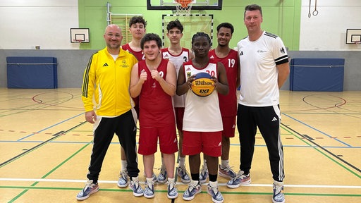 Fünf junge Basketballspieler stehen mit zwei Trainern vor einem Basketballkorb