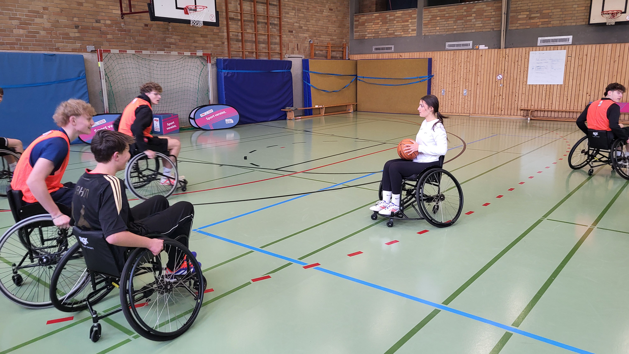 Jugendliche im Rollstuhl spielen in einer Sporthalle Basketball.