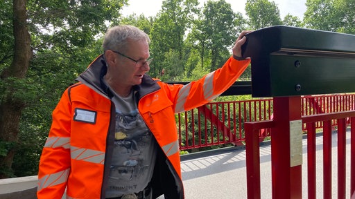 Auf dem Foto ist ein Mann in leuchtend-orangener Baustellenjacke, der am Brückengeländer lehnt.
