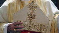 Nahaufnahme von einer Bischofsmütze die gehalten wird. Sie ist mit goldenen Garn und Ornamenten verziert.