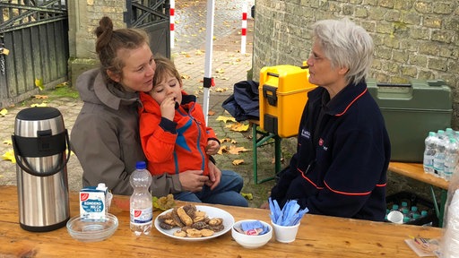 Zwei Frauen sitzen an einem Tisch und unterhalten sich, eine von ihnen hat ein Kind auf dem Schoß sitzen. 