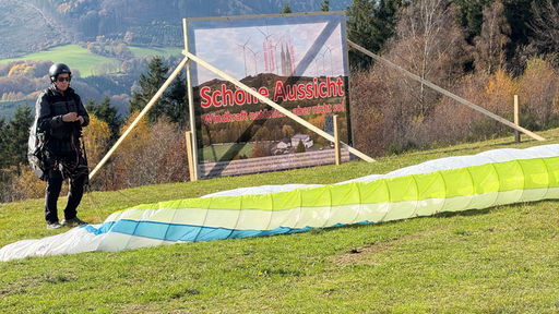 Ein Gleitschirmflieger steht auf einer Wiese. Dahinter ein Plakat auf dem "Schöne Aussicht. Windkraft natürlich aber nicht so!" steht.
