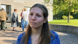Eine Frau steht vor einem Gebäude. Im Hintergrund sind Menschen zu sehen. 