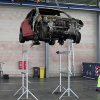 Ein abgeschlepptes E-Auto wird in einer Werkstatt hochgefahren und von zwei Sicherheitsmenschen begutachtet.