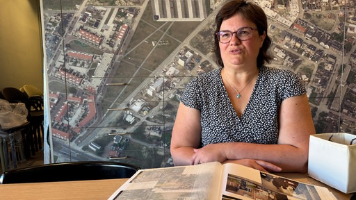 Eine Frau mit Brille sitzt an einem Tisch mit einem aufgeschlagenen Fotoalbum vor einem großen Luftbild an der Wand.