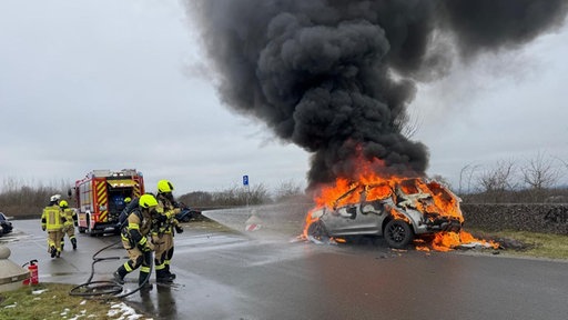 Bild von einem brennenden Auto auf einer Straße, drumherum Feuerwehrkräfte
