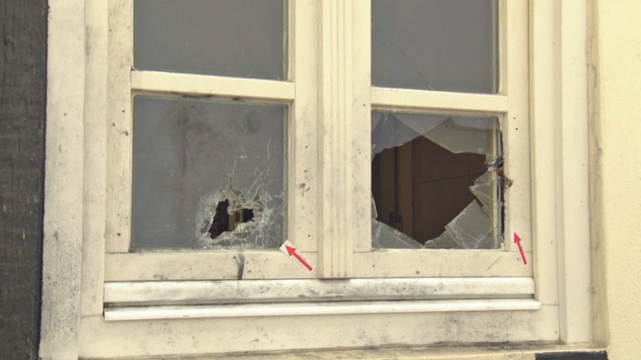 An einem Fenster mit Sprossen sind rote Pfeile angebracht, die auf die unteren Scheiben verweisen, welche Einschusslöcher haben.