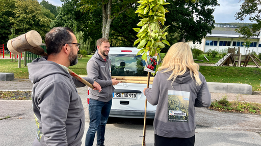 Mehere Personen stehen mit einem jungen Baum vor einem Auto.