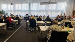 Personen sitzen in einem Sitzungsraum. Eine Person spricht am Sprechpult.