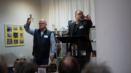 Zwei Männer stehen auf einer Bühne. Einer hält ein Objekt aus Glas hoch. Der andere deutet mit dem Finger in die Menschenmenge vor ihnen.