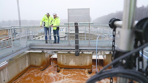 Zwei Männer mit weißem Schutzhelm und grüner Warnkleidung stehen auf einem Metallsteg am Geländer und schauen auf braunes Wasser hinab.