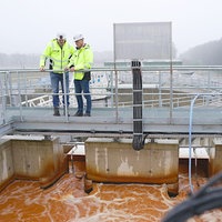 Zwei Männer mit weißem Schutzhelm und grüner Warnkleidung stehen auf einem Metallsteg am Geländer und schauen auf braunes Wasser hinab.