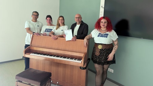 Die Musikerinnen und Musiker stehen gemeinsam mit dem Oberstufenleiter der Gesamtschule hinter einem Klavier