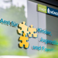 Die Eingangstür des Jugendamts in Münster. Darauf steht "Amt für Kinder, Jugendliche und Familie" dazwischen drei Puzzleteile.