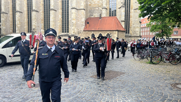 Mehrere Männer in Uniformen und mit Fahnen schreiten über eine Straße