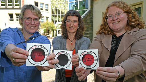 Drei Frauen die Sticker mit der Aufschrift "Hitze Warner" ins Bild halten.