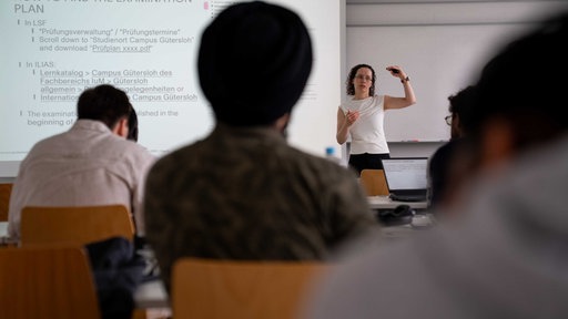 Ein Seminarraum, in dem eine Dozentin vorn einen Vortrag hält. Davor sitzen Studierende, die ihr zuhören.
