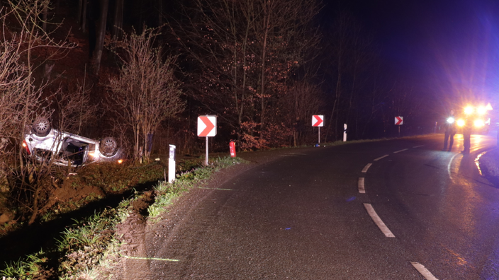 Ein Auto liegt demoliert und auf dem Dach in einem Waldstück neben der Straße. Es sind Reifenspuren zu sehen die von der Straße weggehen in richtung Waldstück. 