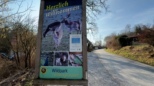 Ein Schild an einer Straße. Auf dem Schild sieht man Rehe und Hirsche und die Aufschrift "Herzlich Wilkommen - WildparkVöllinghausen".