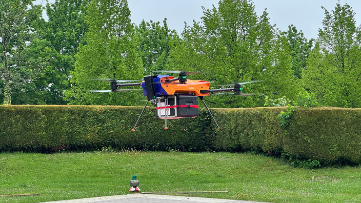 Eine Drohne die fliegt. Im Hintergrund sind eine Wiese, Bäume und eine Hecke zu sehen. 
