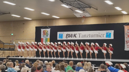 Das Bild zeigt die Tänzerinnen und Tänzer in einer Reihe stehend und dabei synchron das Bein in der Luft.