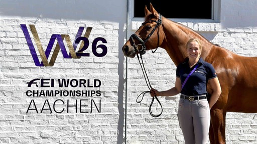 Katharina Hemmer steht mit ihrem Pferd Denoix vor einer weißen Wand auf dem das Logo der WM zu erkennen ist.