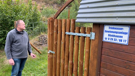 Ein Mann steht vor einem Holztor. Auf einer Wand daneben steht "Trinkwasser-Gewinnungsanlage Bönkhausen"