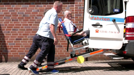 Patient wird vor einer braunen Backsteinwand über eine Rampe in ein Krankentaxi geschoben
