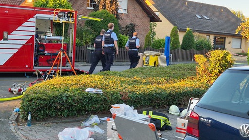 Polizeibeamte gehen auf ein Wohnhaus zu, links im Bild sieht man ein Einsatzfahrzeug der Feuerwehr und im Vordergrund liegt Ausrüstung der Feuerwehr