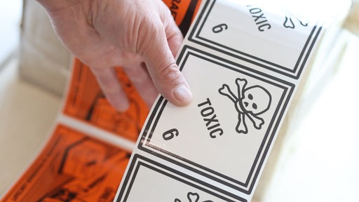 Eine Hand hält Aufkleber mit der Aufschrift "Toxic" und einem Totenkopfbild darauf.
