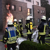 Mehrere Feuerwehrleute stehen vor dem brennenden Haus und schauen auf eine Stichflamme, die gerade aus einem Kellerfenster kommt und an der Fassade empor steigt