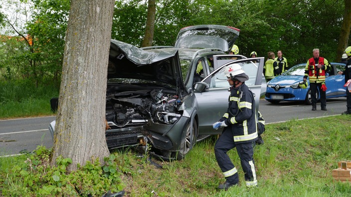 Ein völlig zerstörtes Auto, das gegen einen Baum gefahren ist. Im Hintergrund Rettungskräfte.
