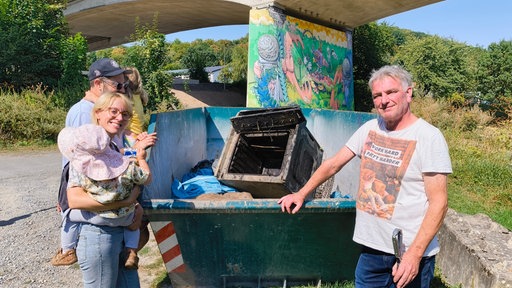 Drei Personen stehen vor einer Mulde. In der Mulde befindet sich, auf einem Schrotthaufen, ein kaputter, rostiger Backofen ohne Innenleben.