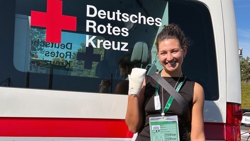 Eine Frau steht vor einem Wagen des deutschen roten Kreuzes mit einem Verband und lächelt in die Kamera.